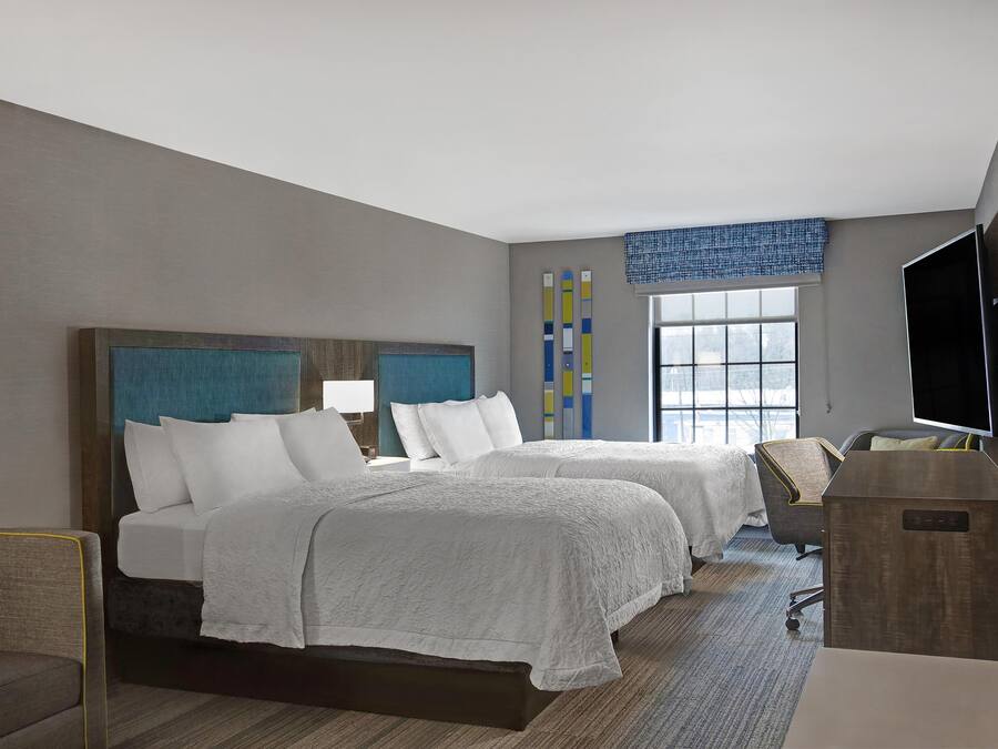 A hotel room with two beds, a desk, a TV, and a window with blinds.