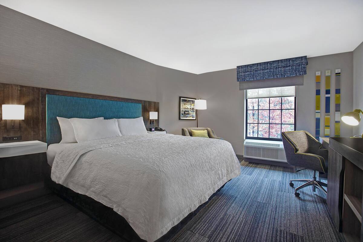 A hotel room with a large bed, a desk with a chair, a window with a view, and a wall-mounted lamp.