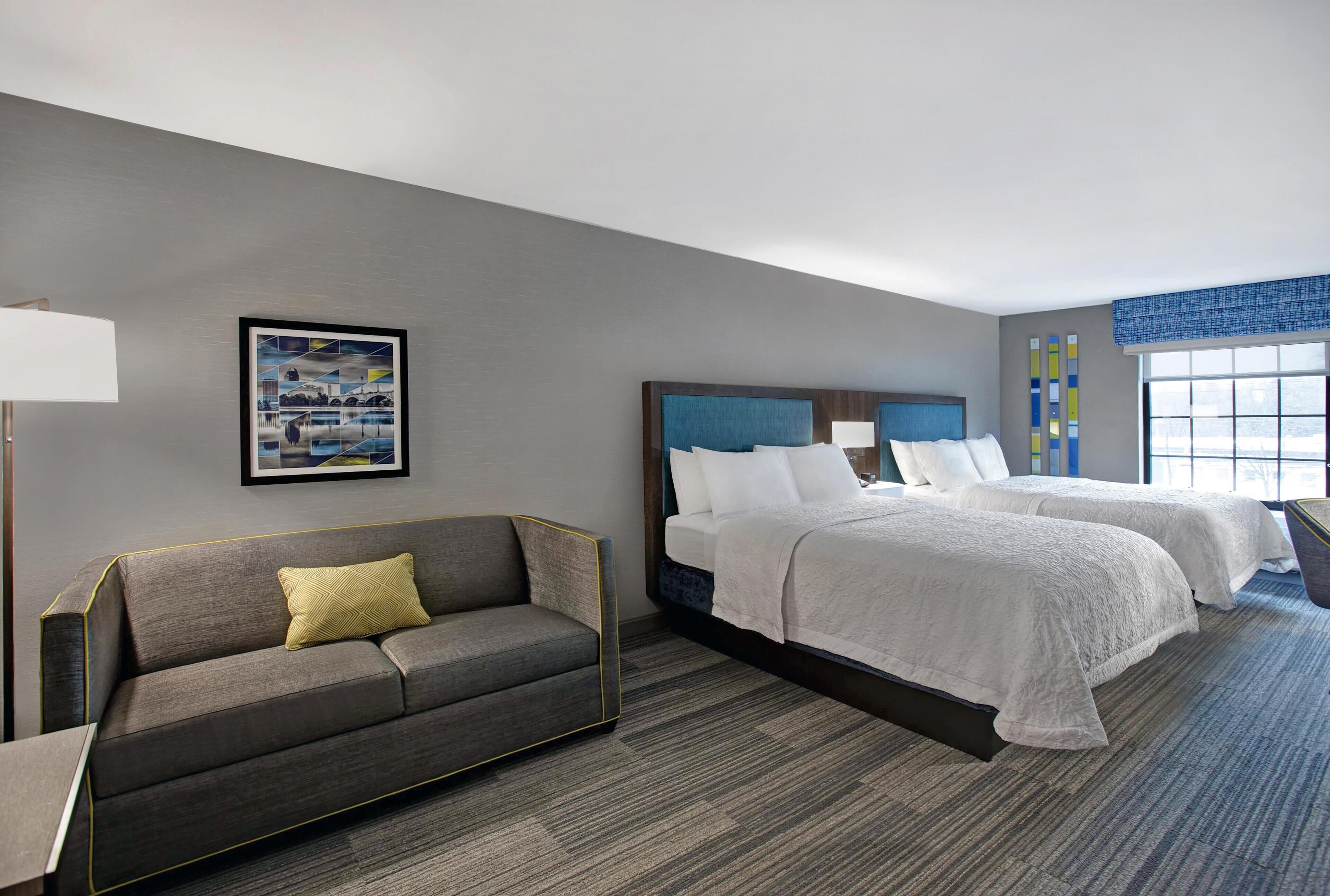 Een hotelkamer met een grijze bank, twee bedden met witte lakens, een ingelijst schilderij aan de muur en een raam met blauwe jaloezieën.