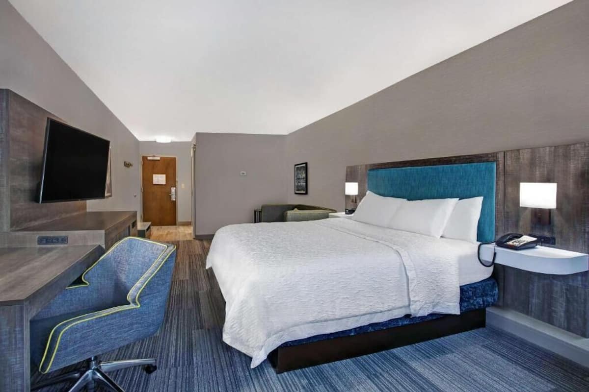 A hotel room with a bed, a desk, a chair, a TV, and a bedside table.