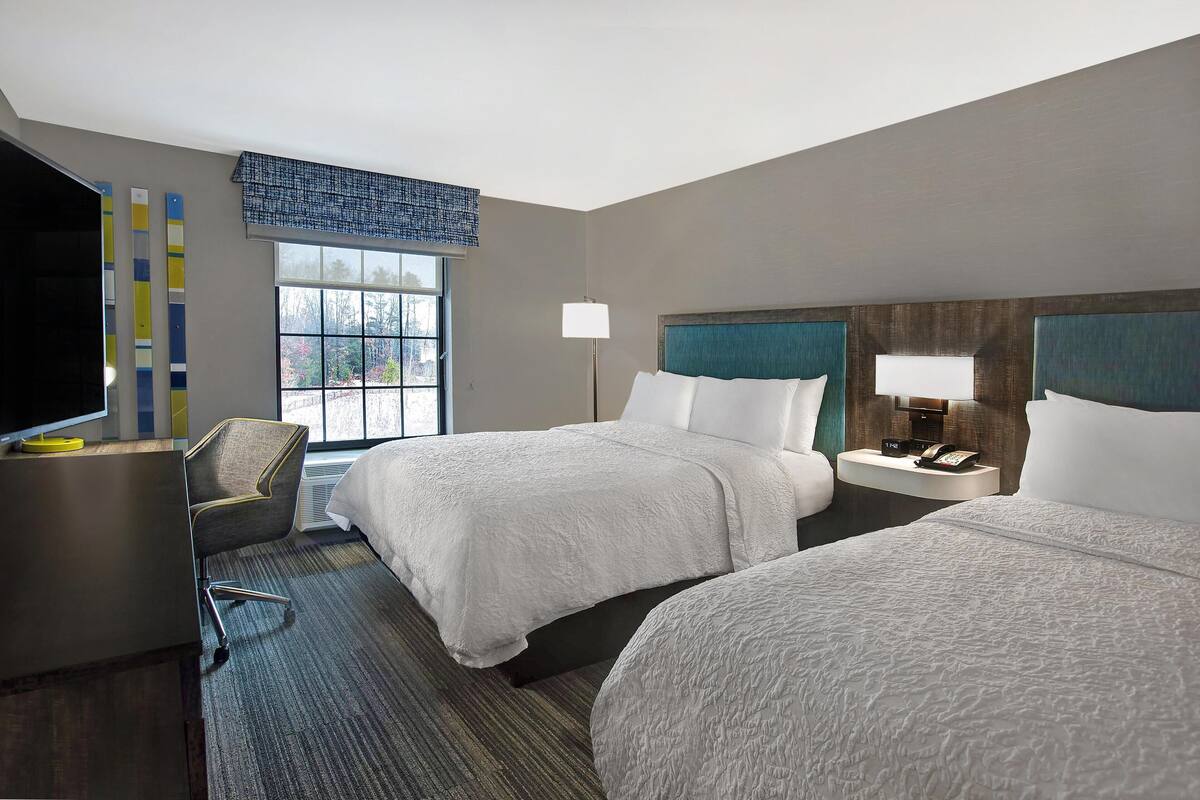 A hotel room with two beds, a desk, a chair, a TV, and a window with a view of trees.