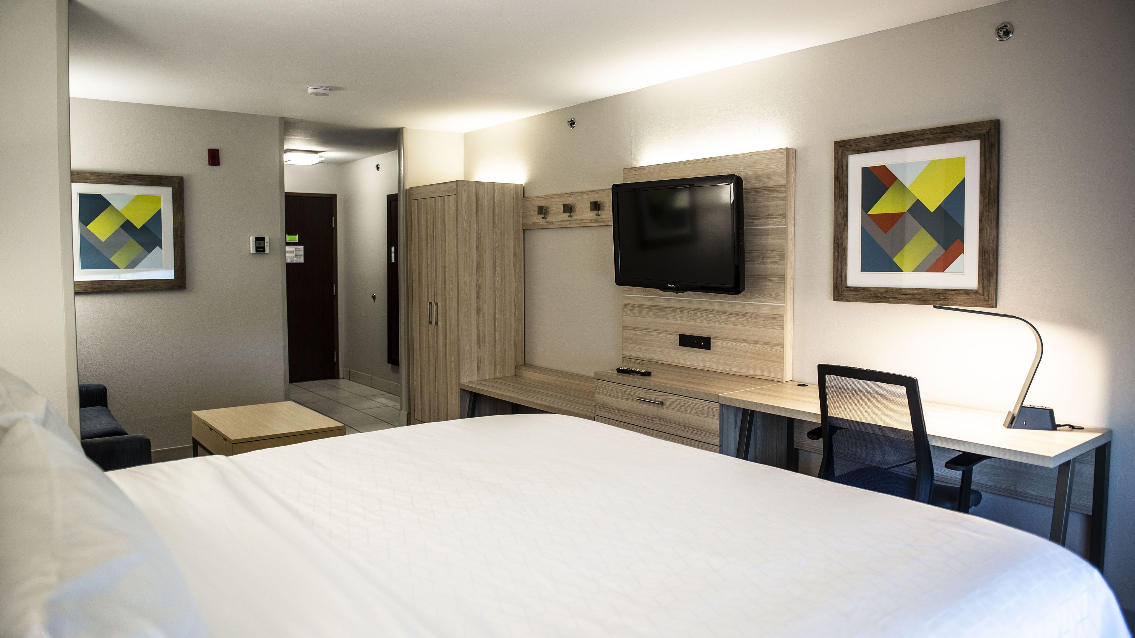 A hotel room with a large bed, a desk with a chair, a TV mounted on the wall, and artwork on the walls.