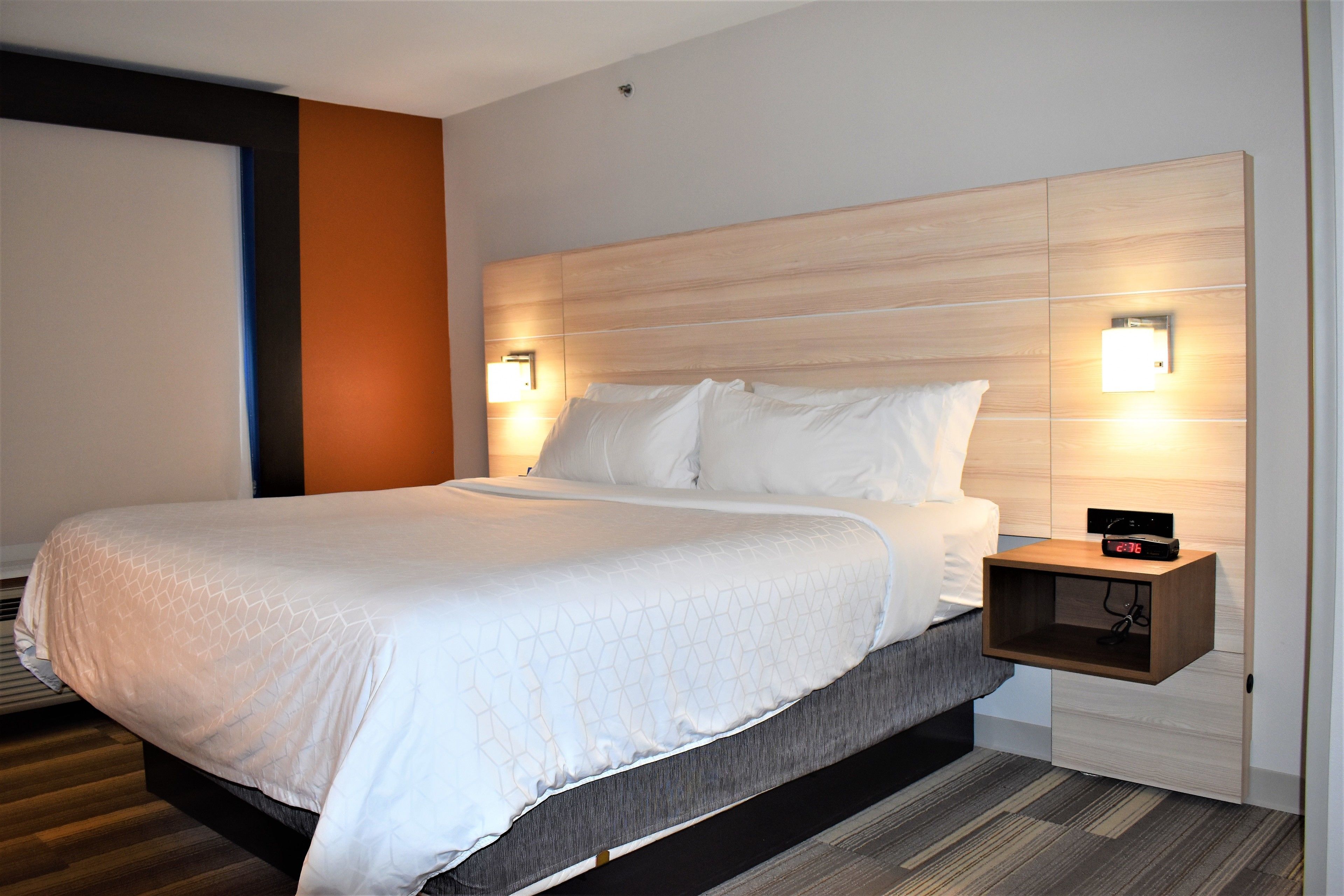 Una habitación de hotel moderna con una cama grande, un cabecero de madera, dos lámparas de noche y una mesita de noche con un reloj.