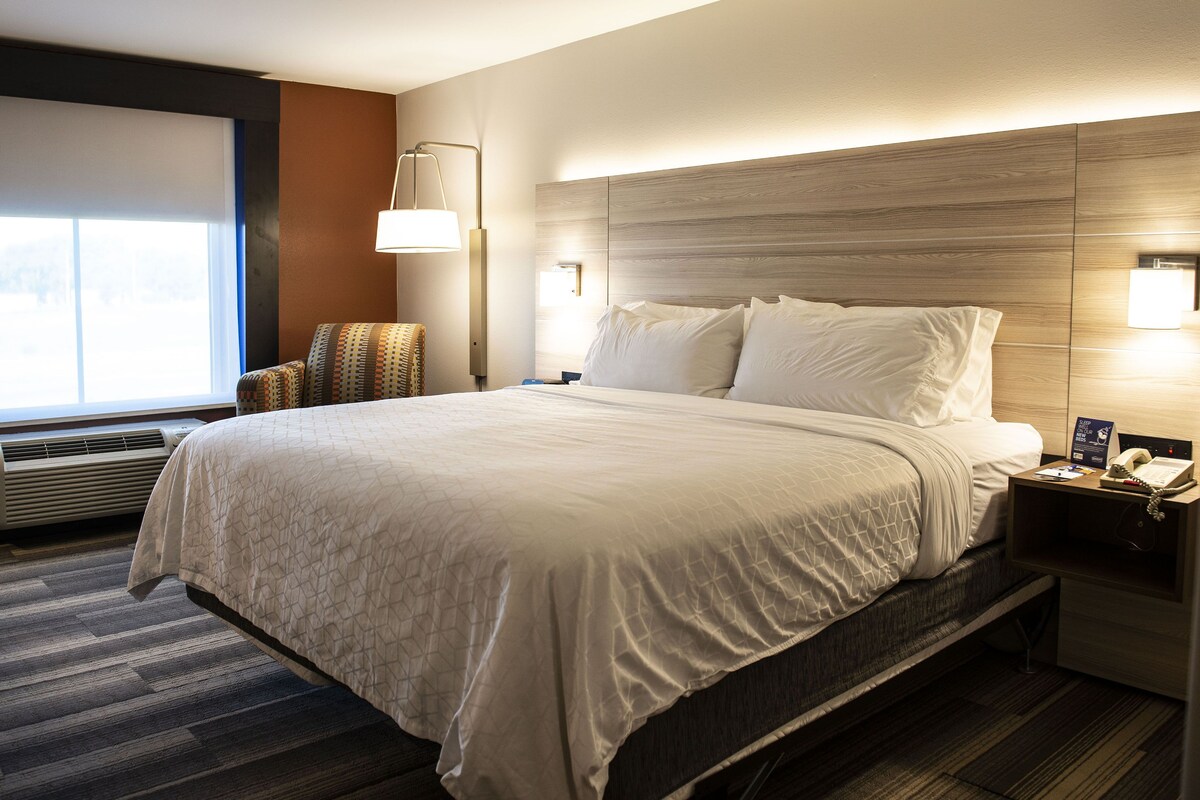 A hotel room with a large bed, a bedside table with a lamp, a nightstand with a phone, and a window with blinds.