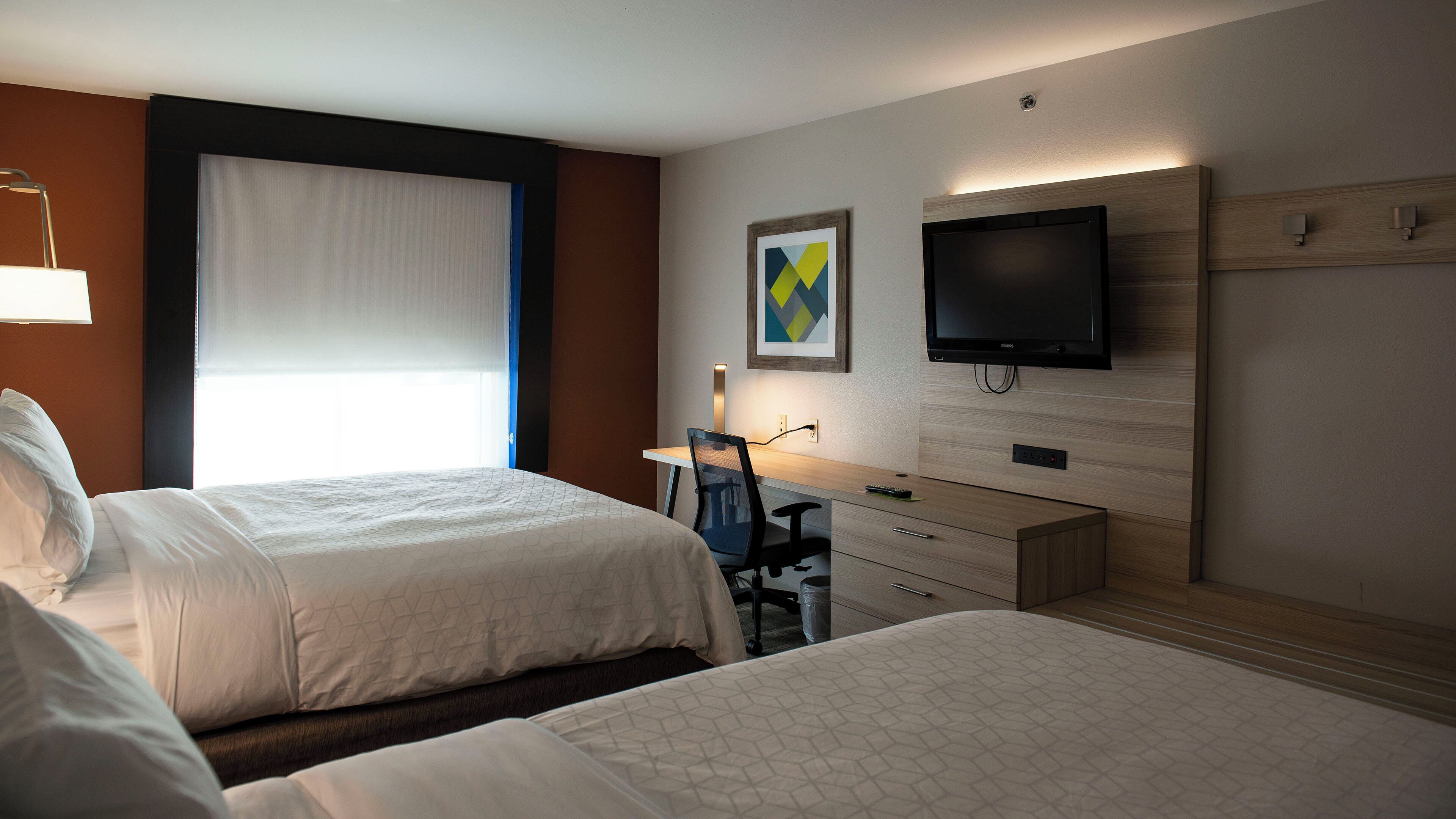 Ein Hotelzimmer mit zwei Betten, einem Schreibtisch mit Stuhl, einem Fernseher und einem Fenster mit Jalousien.
