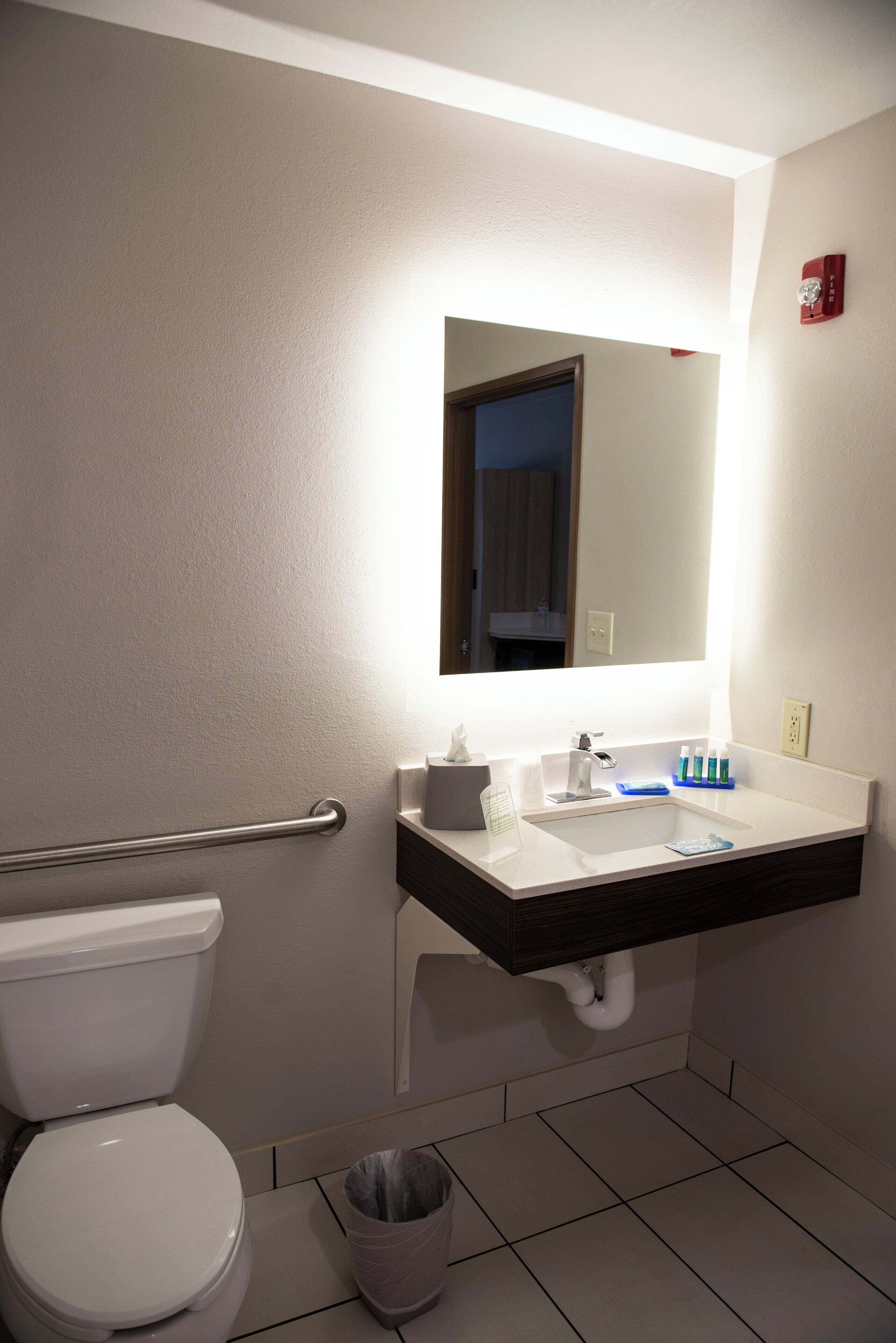 Une salle de bain équipée de toilettes, d’un lavabo, d’un miroir et d’une barre d’appui.