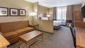 Een hotelkamer met een sofa, een tafeltje, een bed, een bureau en een televisie. Er hangen foto's in lijsten aan de muur en er is een raam met gordijnen.