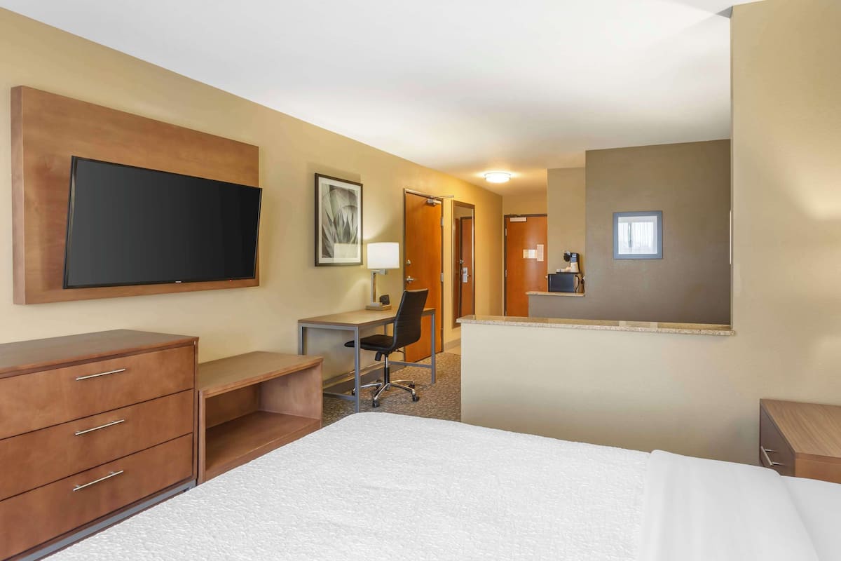 A hotel room with a bed, a desk with a chair, a television mounted on the wall, and a door leading to another room.