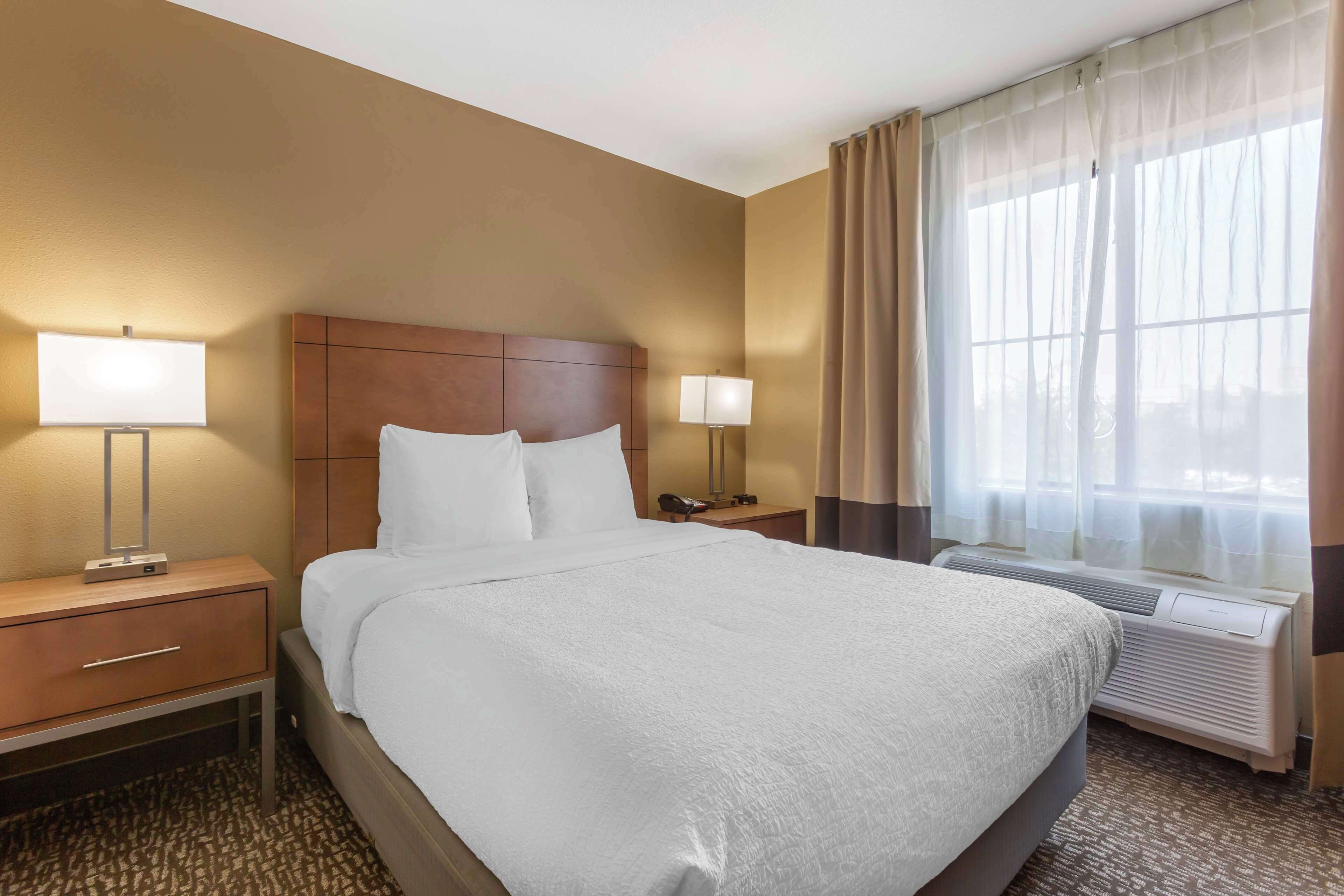 A hotel room with a large bed, two bedside tables with lamps, a headboard, an air conditioning unit, and a window with sheer curtains.