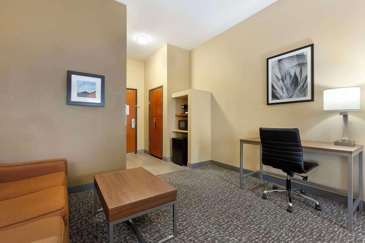 A hotel room with a wooden desk, black office chair, and a small table. There is a framed picture on the wall and a lamp on the desk.
