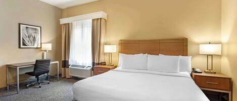 Een hotelkamer met een groot bed, een bureau met stoel, twee nachtkastjes met lampen, een raam met gordijnen en een wandairconditioning.
