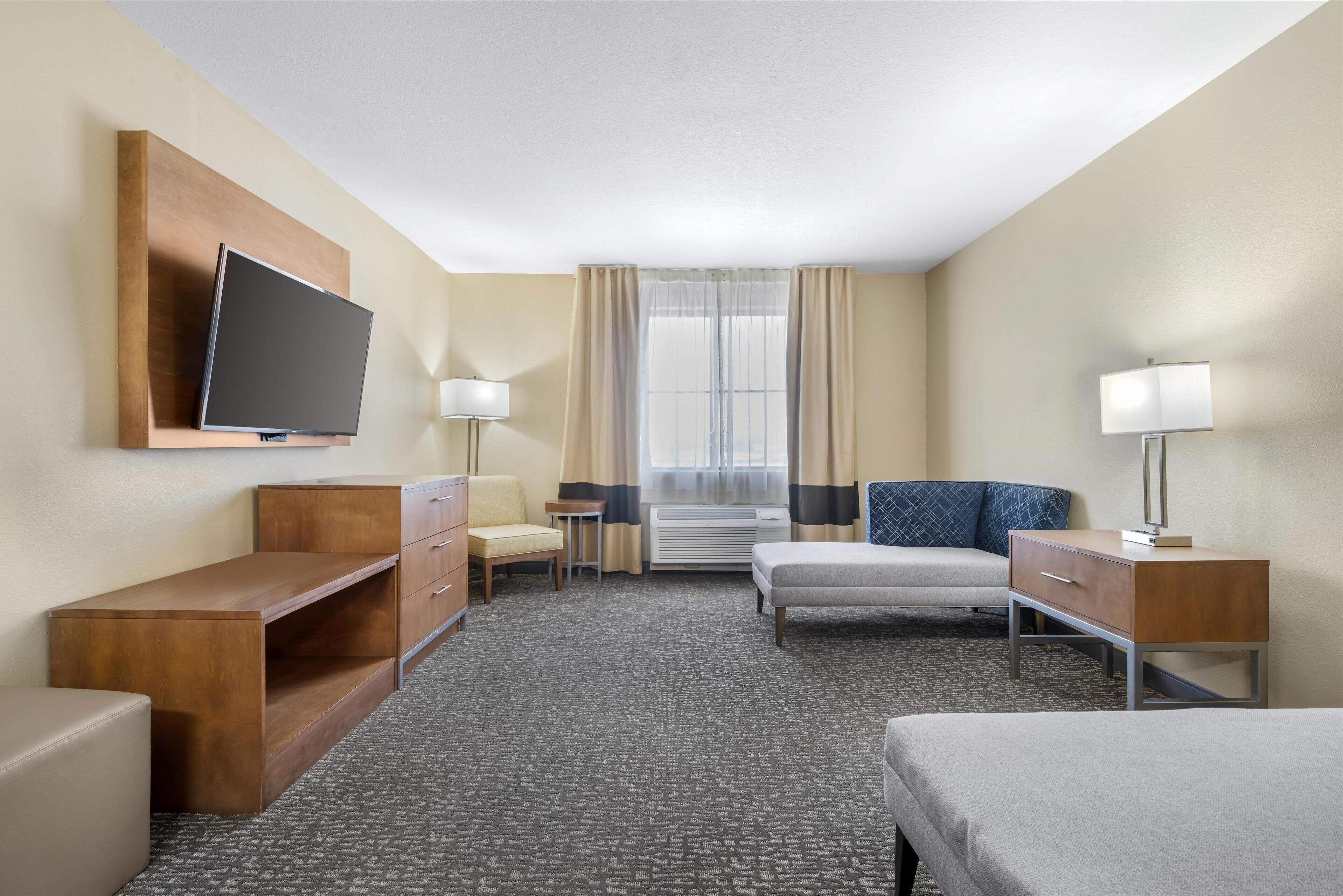 A hotel room with a flat-screen TV, a desk, a chair, a bed, a bedside table, a lamp, and a window with curtains.