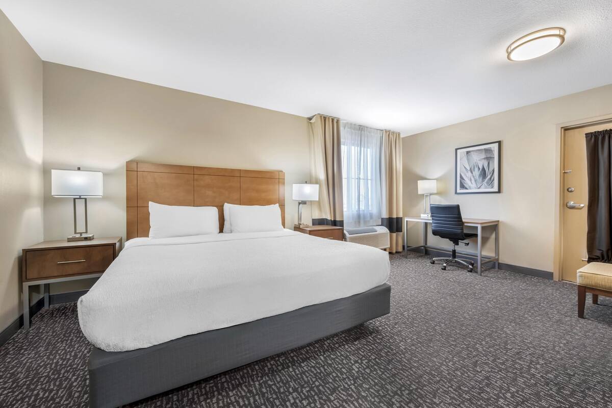 A hotel room with a large bed, two bedside tables with lamps, a desk with a chair, a window with curtains, and a framed picture on the wall.