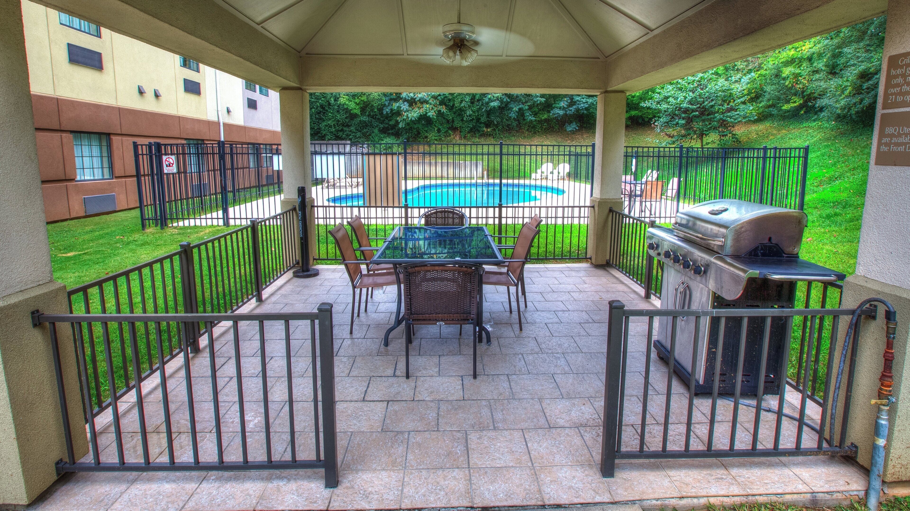 Une terrasse couverte avec une table et des chaises, un barbecue et une vue sur une piscine et la verdure.