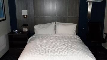 Habitación de hotel con una cama grande, cabecero, mesita de noche con lámpara y un reloj de pared.