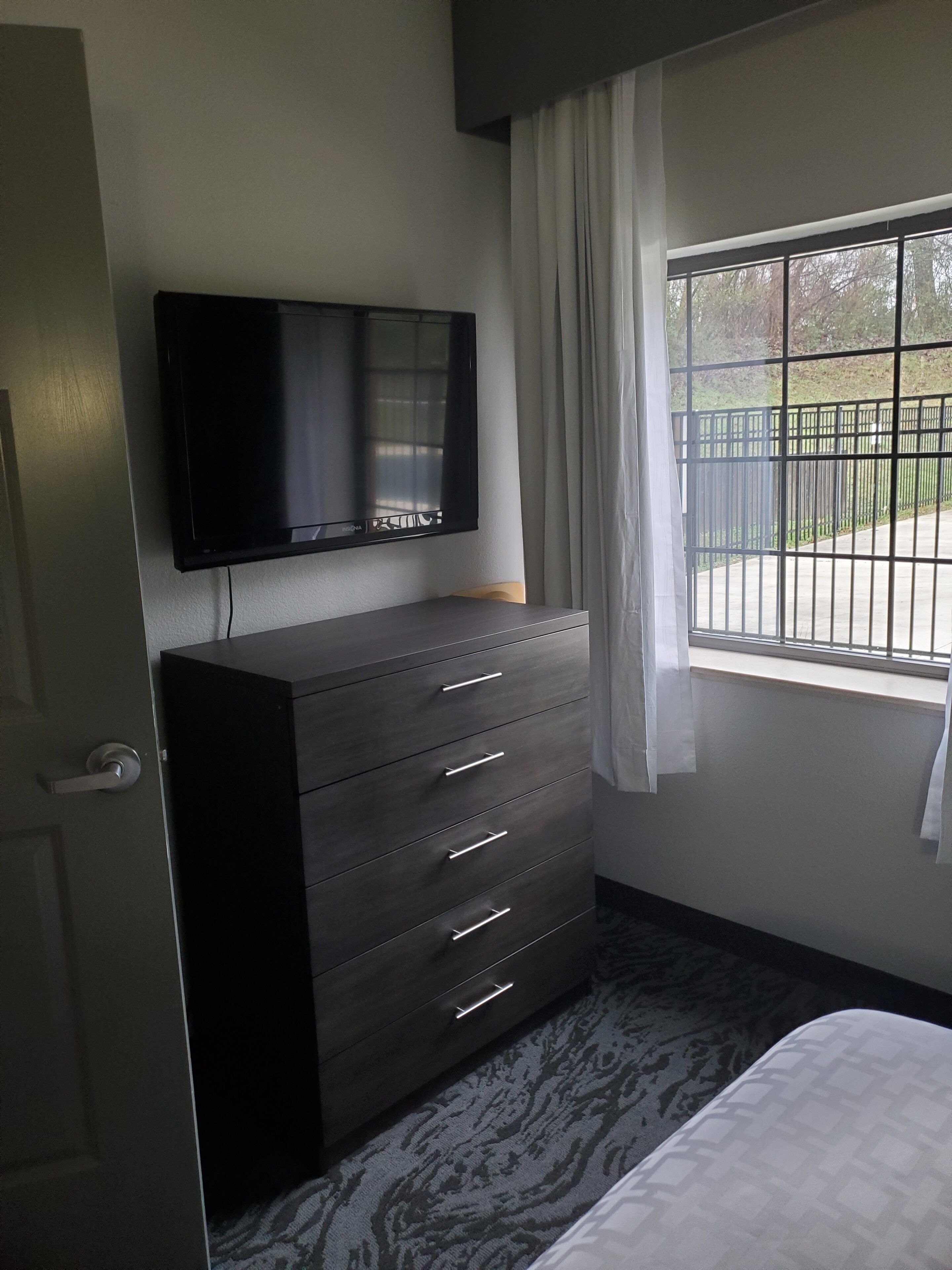 Un dormitorio con un aparador de madera oscura, un televisor de pantalla plana montado encima y una ventana que ofrece vistas a una cerca y vegetación.