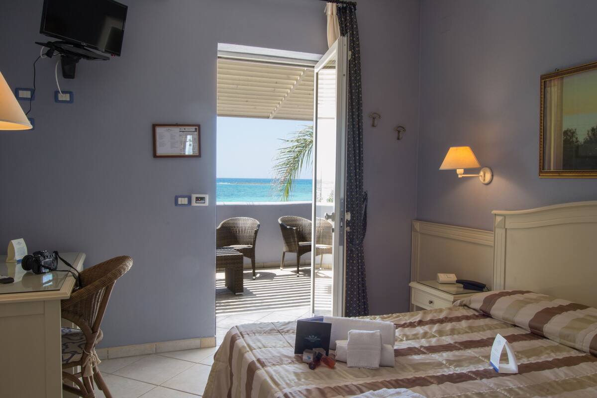 Habitación de hotel con una cama, un escritorio con una cámara, una silla, una mesita de noche y vista al océano.