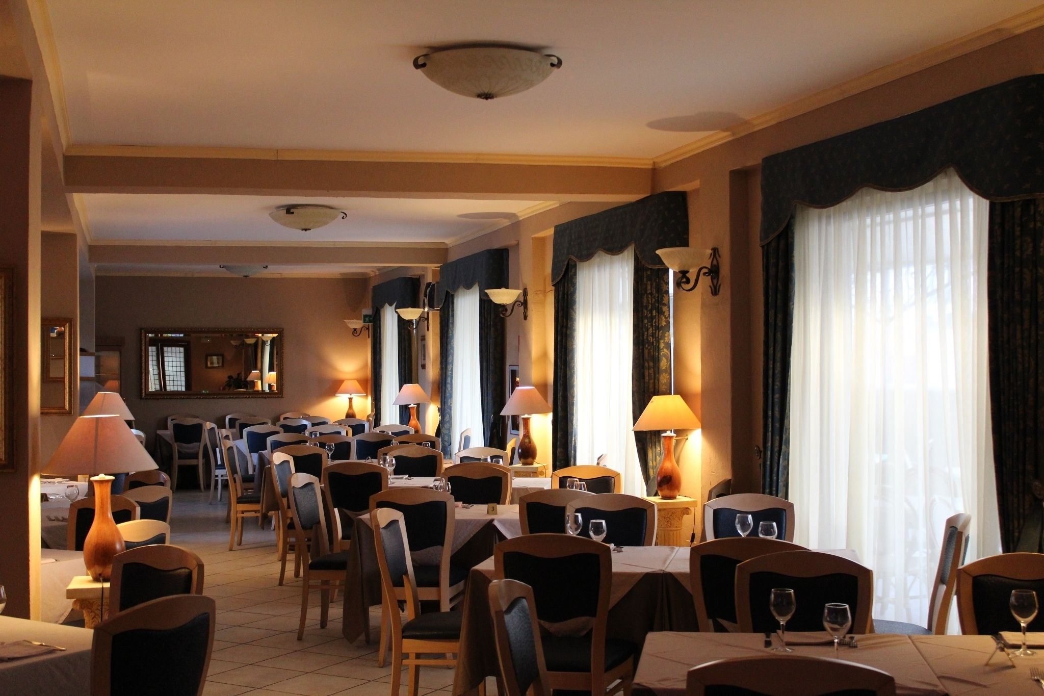 Ein Hotel-Essbereich mit gedeckten Tischen, Deckenleuchten und großen Fenstern mit Vorhängen.