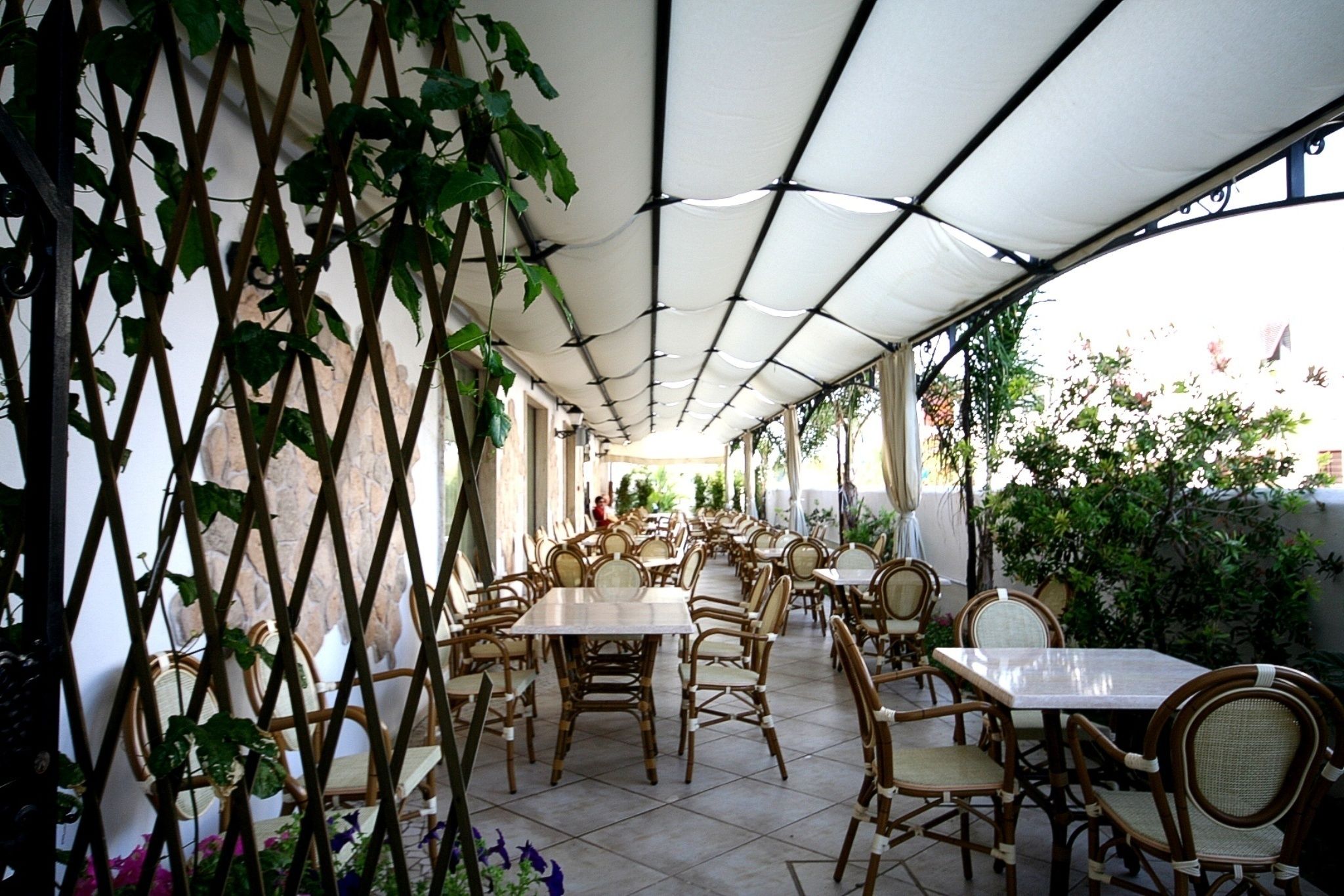 An outdoor café with tables and wicker chairs, covered by a white canopy, and surrounded by greenery.
