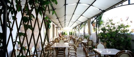 An outdoor café with tables and wicker chairs, covered by a white canopy, and surrounded by greenery.