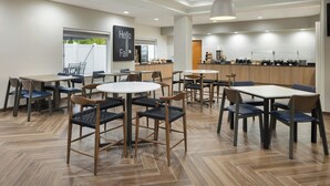 A modern dining area with multiple tables and chairs, a kitchen in the background, and a 'Hello Fall' sign on the wall. - Fairfield Inn & Suites Lake City (Lake City)