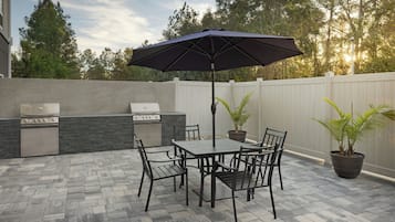 Une terrasse extérieure équipée d’un barbecue, d’une table, de chaises et de plantes en pot.