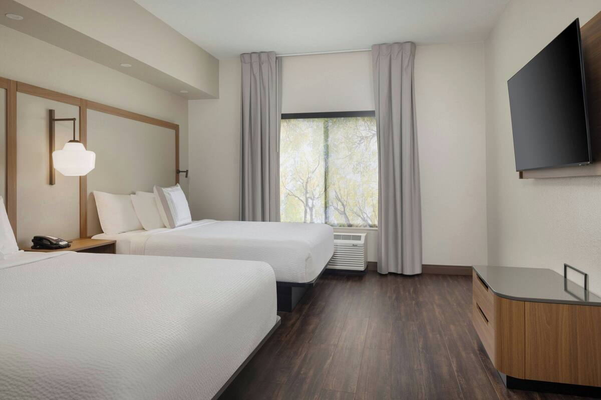 A hotel room with two beds, a flat-screen TV, a bedside table with a lamp, and a large window with curtains.