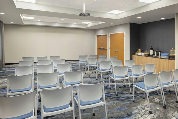 A conference room with rows of white chairs with blue cushions, a podium with water bottles, and a projector on the ceiling.