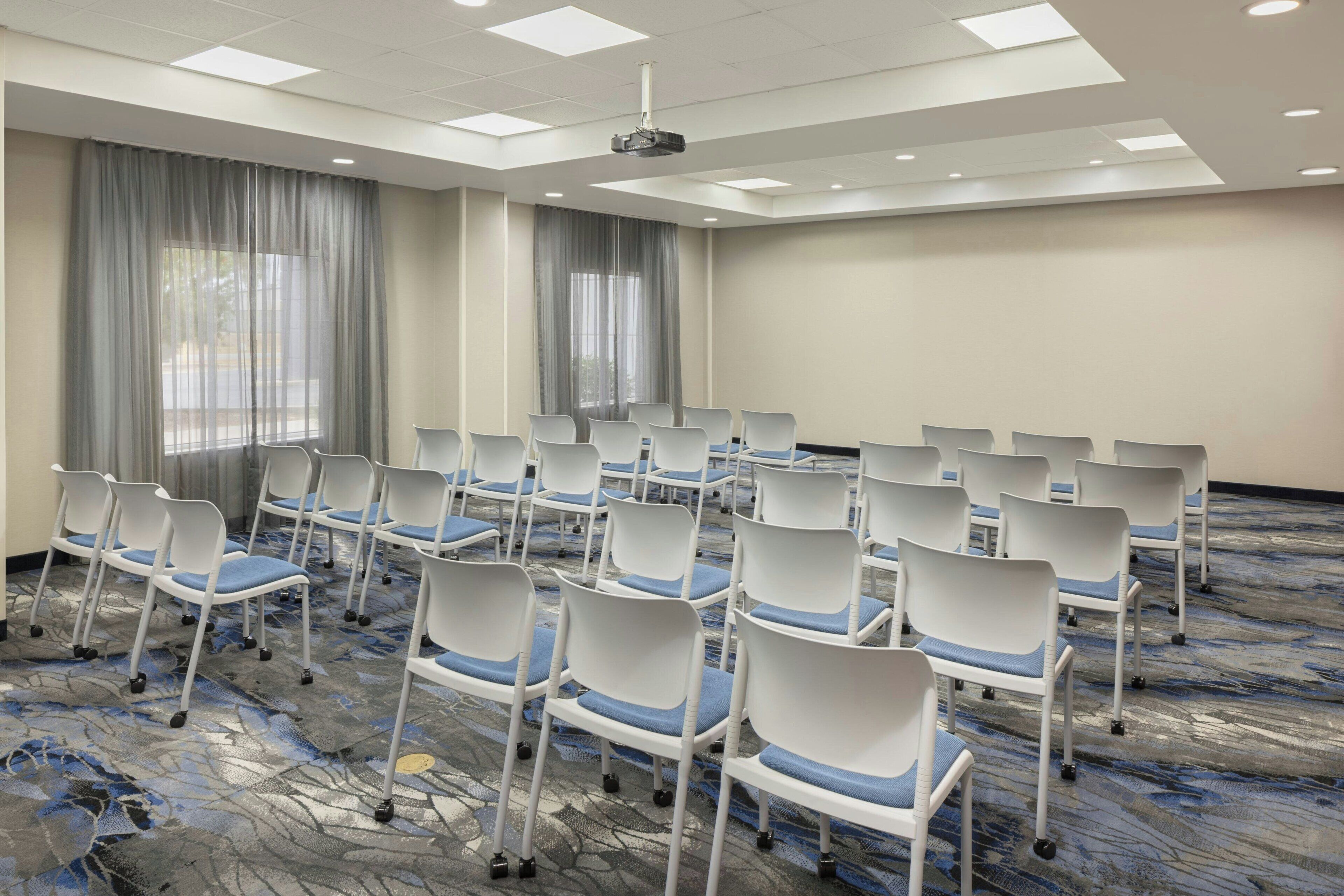 A conference room with rows of white chairs with blue cushions, a projector on the ceiling, and large windows with curtains.
