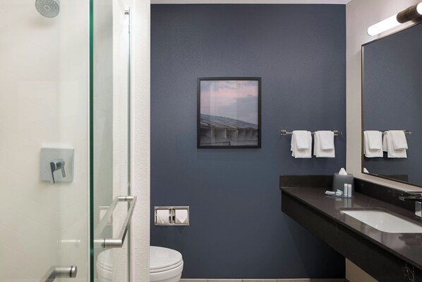 Un baño moderno con cabina de ducha de vidrio, un inodoro blanco, una pared azul oscuro, un cuadro enmarcado y un espejo grande con toallas.