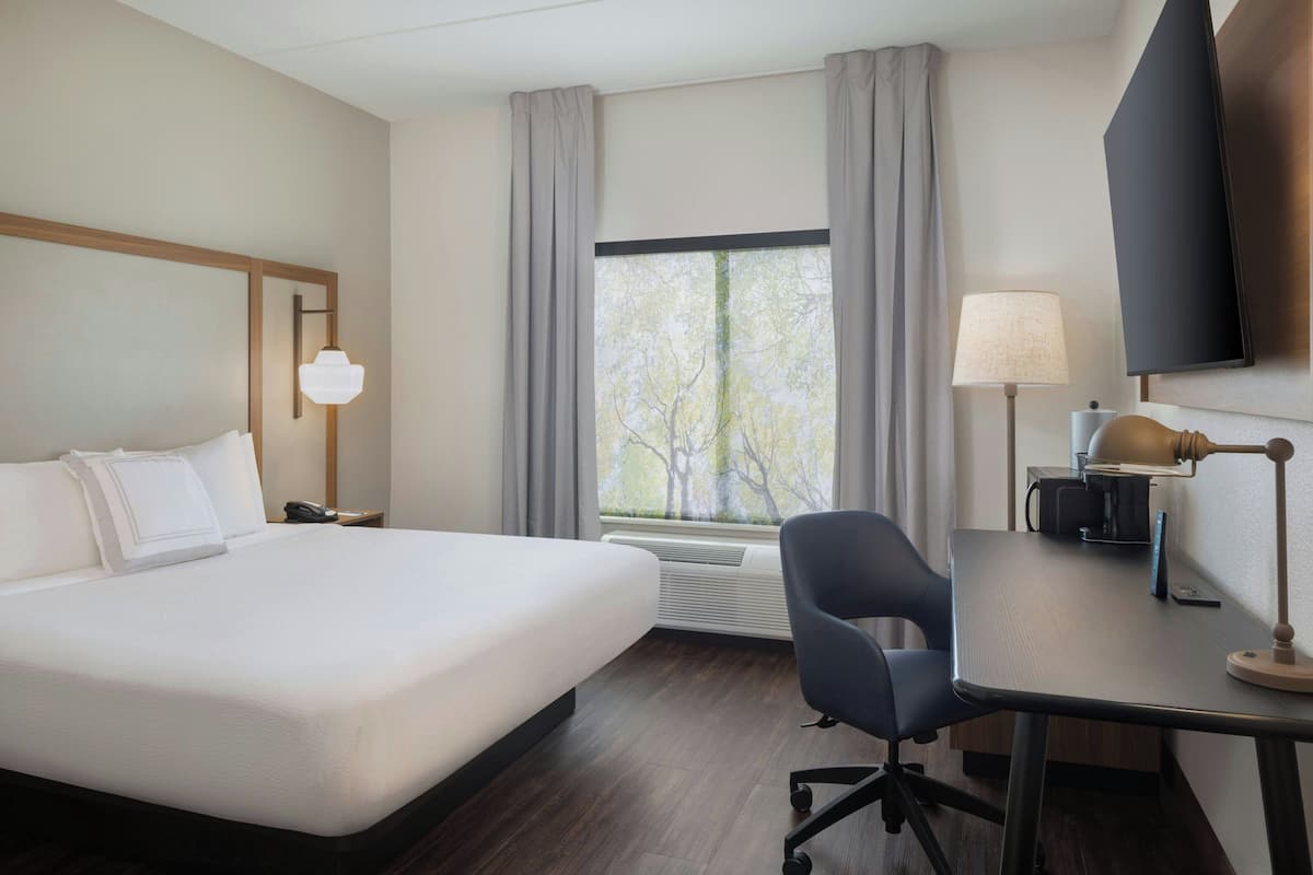 A hotel room with a large bed, a desk with a lamp, a chair, a TV mounted on the wall, and a window with curtains.
