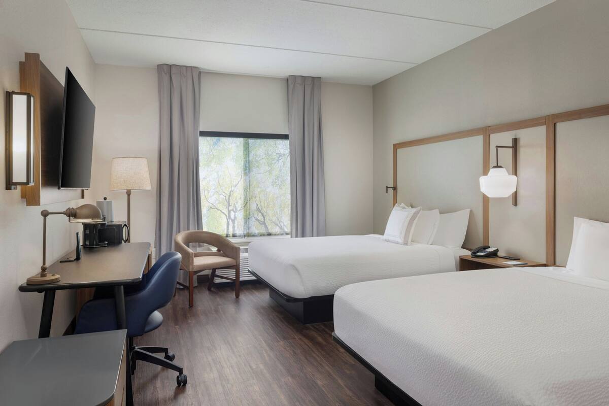 A hotel room with two beds, a desk with a chair, a TV, and a window with curtains.