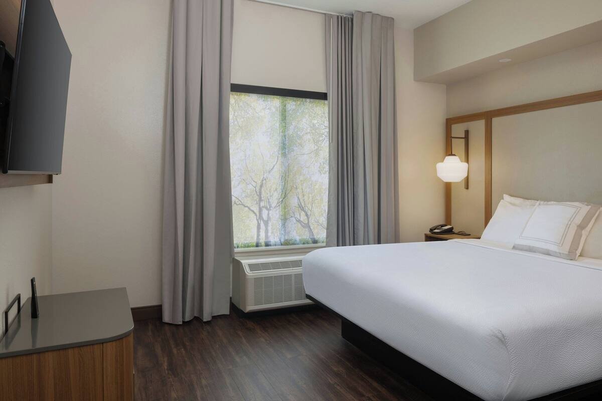 A hotel room with a large bed, a desk, a TV, a window with curtains, and a nightstand with a lamp.