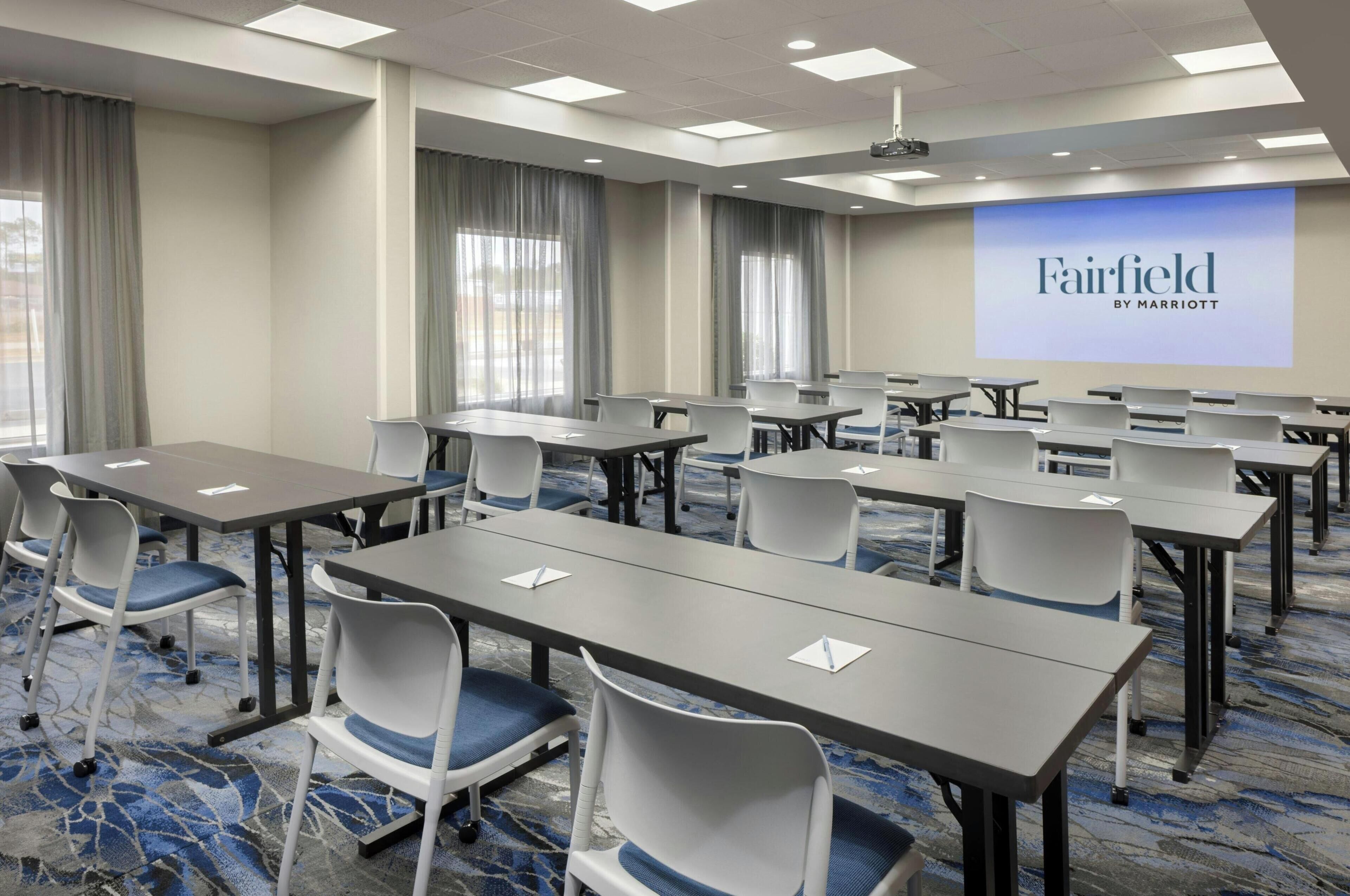 Une salle de conférence dotée de plusieurs tables et chaises, d’un grand écran affichant le logo « Fairfield by Marriott » et d’un sol recouvert de moquette.