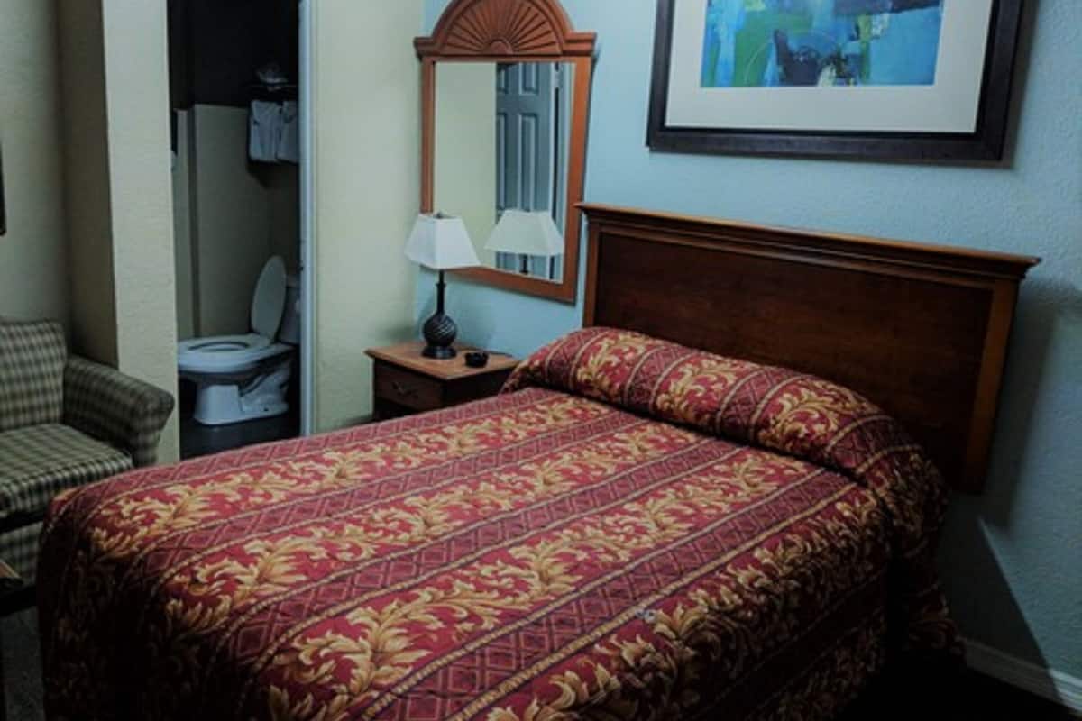 Habitación de hotel con cama, una silla, mesita de noche, espejo y un cuadro en la pared.
