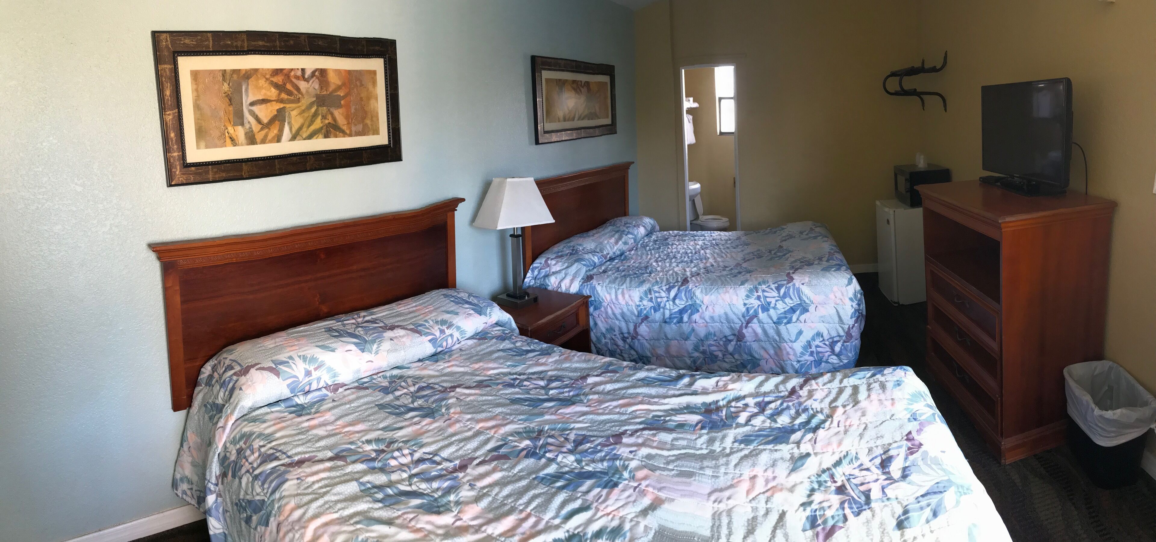 Une chambre d’hôtel avec deux lits, une tête de lit en bois, une table de chevet avec une lampe, une commode, un téléviseur et des œuvres d’art encadrées accrochées au mur.