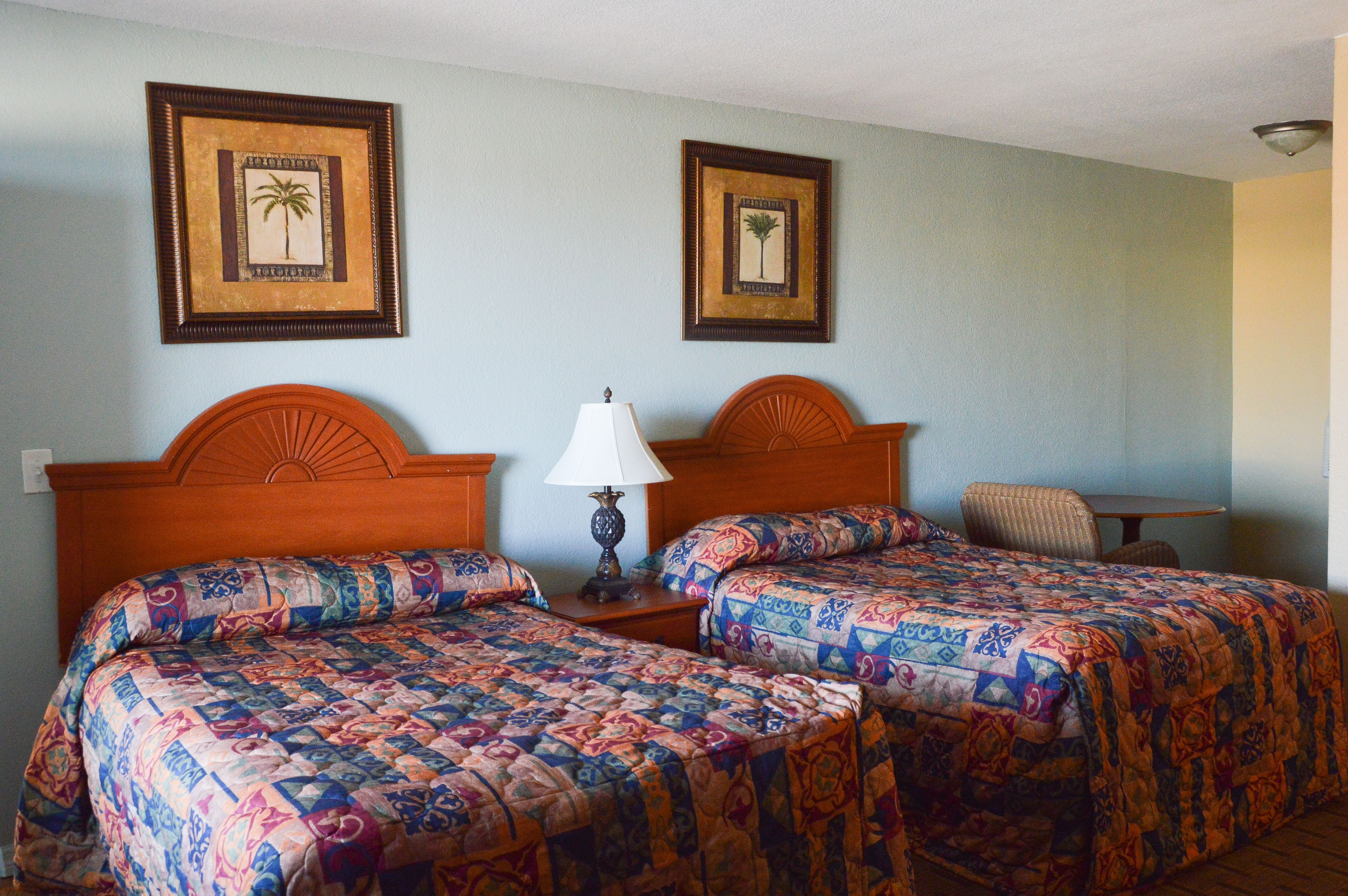 A hotel room with two beds, a nightstand with a lamp, a chair, and two framed pictures on the wall.
