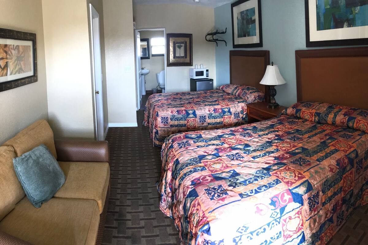 Habitación de hotel con un sofá marrón, dos camas con colchas coloridas, un microondas, un lavamanos y obras de arte enmarcadas en las paredes.