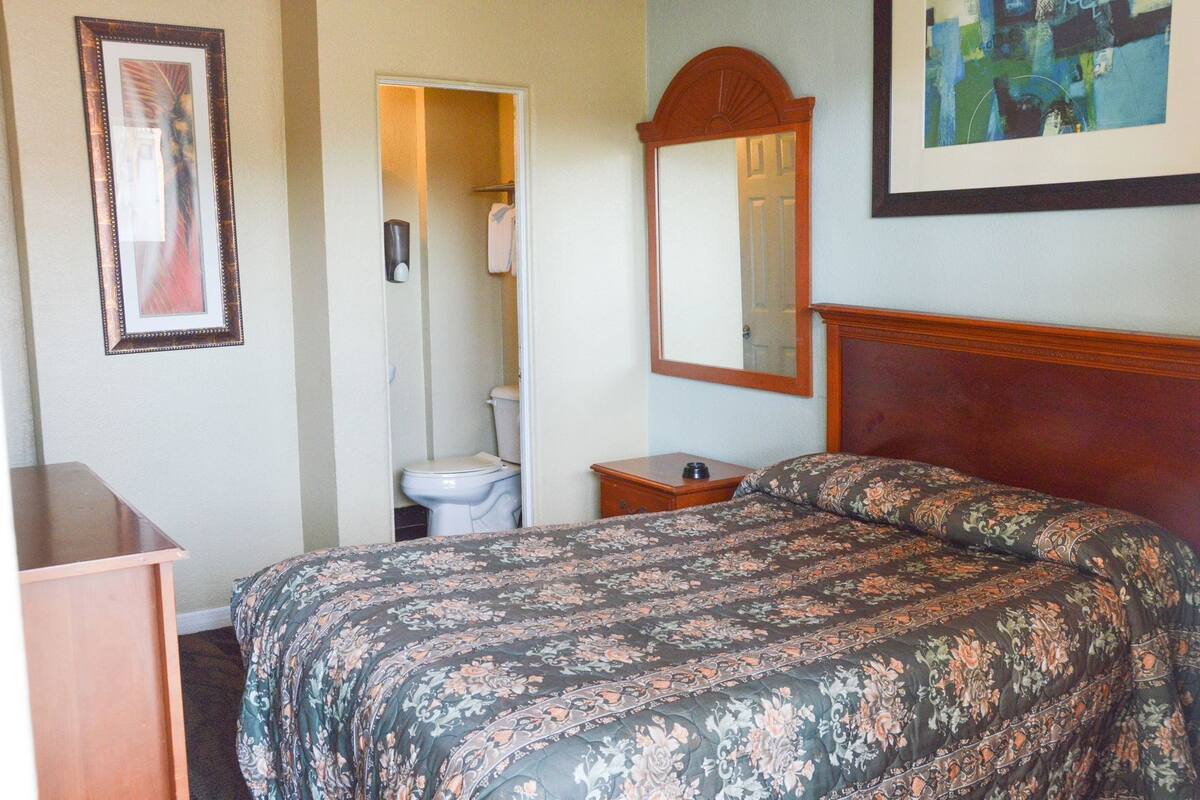 Un dormitorio con cama, cabecera de madera, mesita de noche, espejo, un cuadro en la pared y baño con inodoro y ducha.