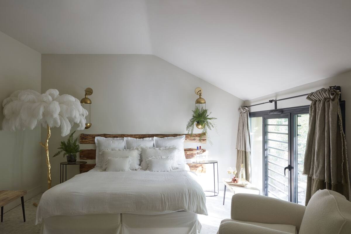 Un dormitorio con una cama grande, un banco, una planta en maceta y una ventana con cortinas.