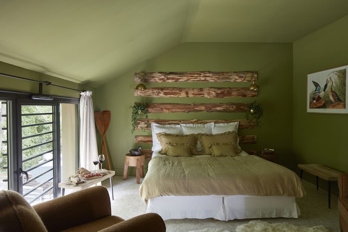 Un dormitorio con una cama, cabecera de madera, una ventana, un banco y una mesa con una planta.