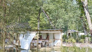 Una cabaña de madera con una pequeña terraza, rodeada de árboles y un estanque.