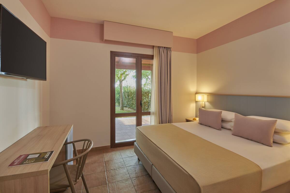 A hotel room with a bed, a desk, a chair, a TV, and a window with a view of trees.