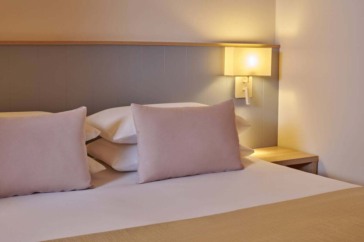 A hotel room with a neatly made bed, a bedside table, and a wall-mounted lamp.