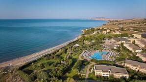 An aerial view of a coastal resort with a beach, a swimming pool, and residential buildings. - Serenusa Resort (Licata)