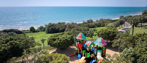 A playground with slides and climbing structures, surrounded by greenery and a view of the sea.