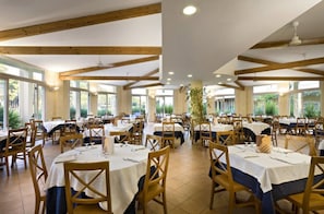 A dining area with multiple tables and chairs, wooden beams on the ceiling, and large windows allowing natural light.