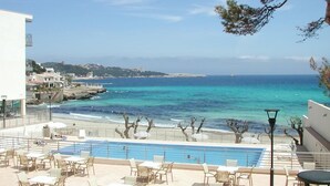 A poolside area with a view of the sea, outdoor seating, and a building in the background. - Hotel Na Forana (Capdepera)