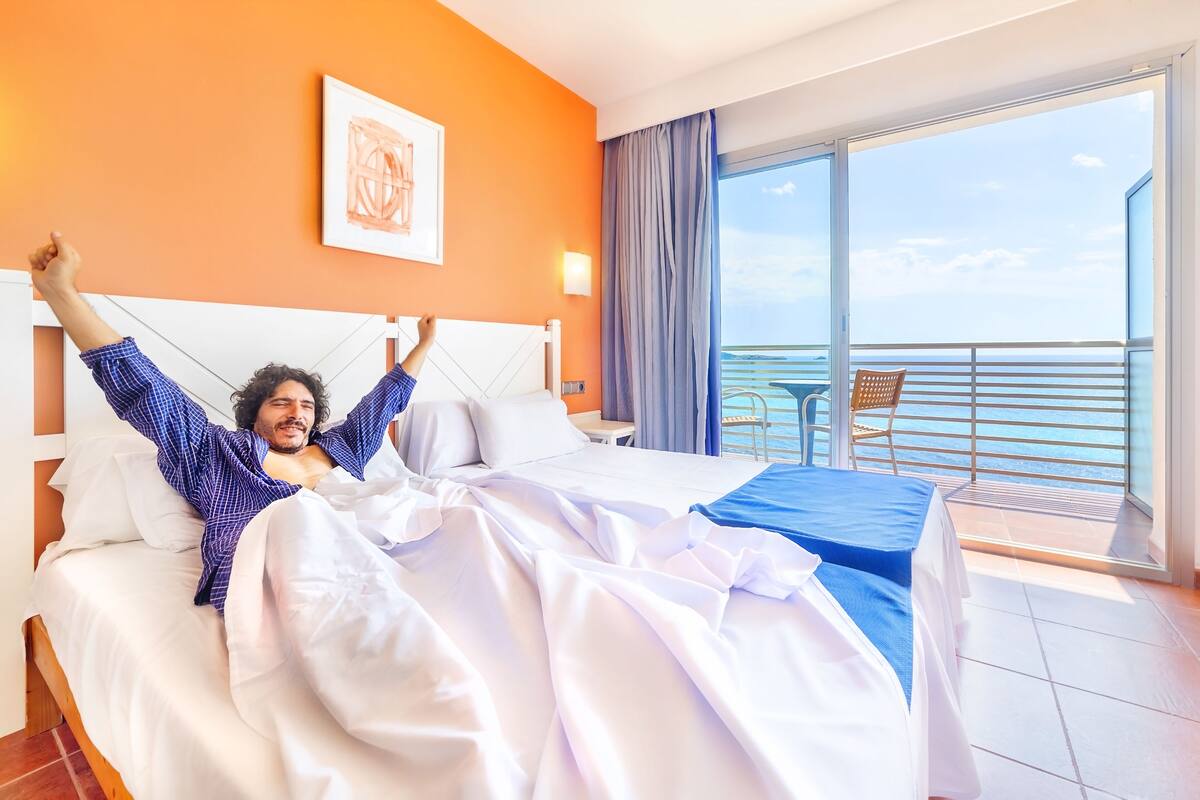 Una habitación de hotel con una cama, un ventanal con vistas al mar y un balcón con mesa y sillas.