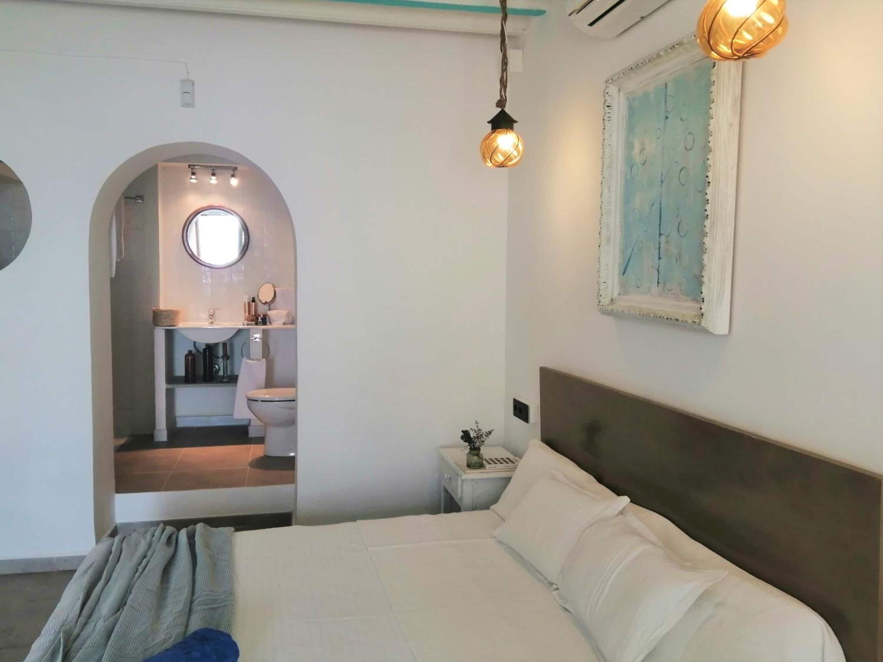 Un dormitorio con una cama, una mesita de noche, una lámpara de pared y un baño visible a través de un arco.